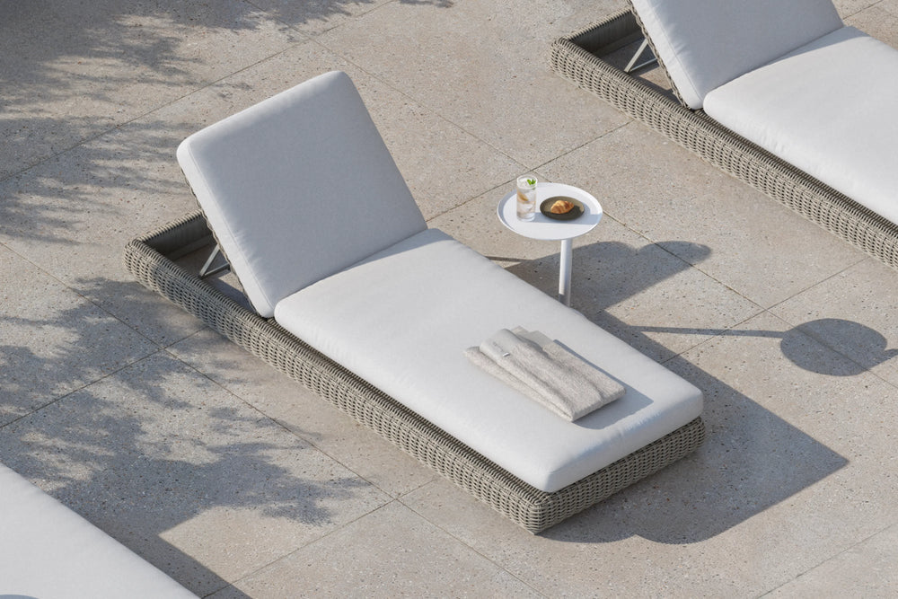 Top-down lifestyle shot of a Neighbor Roundabout Sun Lounger in taupe-gray resin wicker with thick cream cushion on a sunlit stone-tile poolside patio, with a folded towel set on the seat and a small white side table holding lemon water and a croissant beside it.
