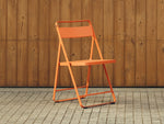 Three-quarter product photo of a single coral-red Neighbor Take Two Bistro Chair — perforated steel back and seat on a slim folding tubular-steel frame — set on a cobblestone patio against a stained-wood plank wall.
