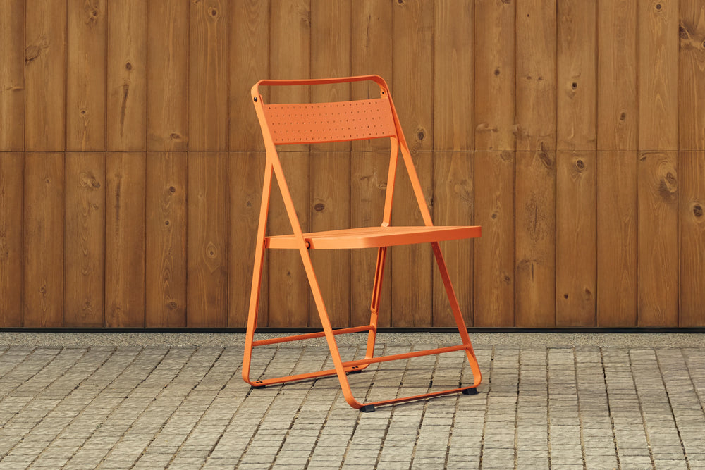 Three-quarter product photo of a single coral-red Neighbor Take Two Bistro Chair — perforated steel back and seat on a slim folding tubular-steel frame — set on a cobblestone patio against a stained-wood plank wall.