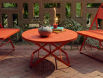 Three-quarter view of a coral-red Neighbor Take Two Coffee Table on a red-brick paver patio styled with a textured stoneware vase, stacked teal and yellow books, and a lit taper candle, flanked by the seats of two coral-red Take Two Lounge Chairs against a cinder-block lattice wall and tropical greenery.