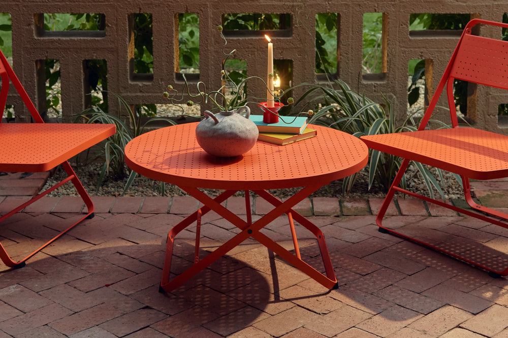 Three-quarter view of a coral-red Neighbor Take Two Coffee Table on a red-brick paver patio styled with a textured stoneware vase, stacked teal and yellow books, and a lit taper candle, flanked by the seats of two coral-red Take Two Lounge Chairs against a cinder-block lattice wall and tropical greenery.
