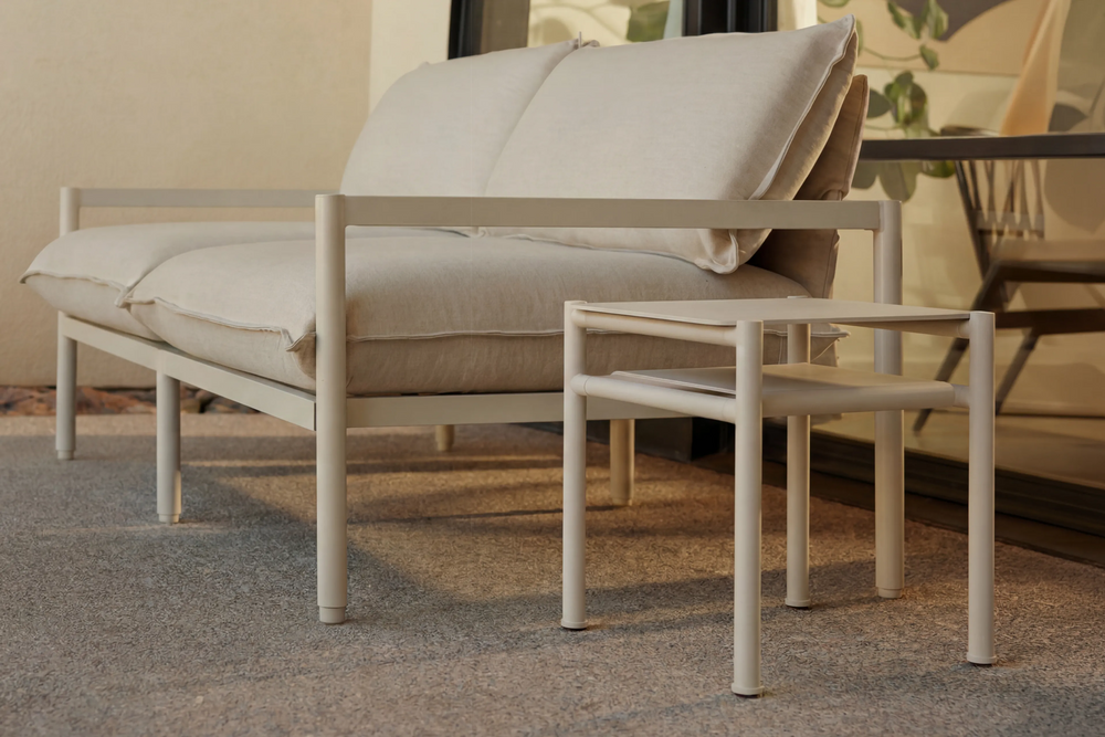 Neighbor Underline Side Table in off-white, a compact two-tier table on slim tubular legs, next to a cushioned sofa.