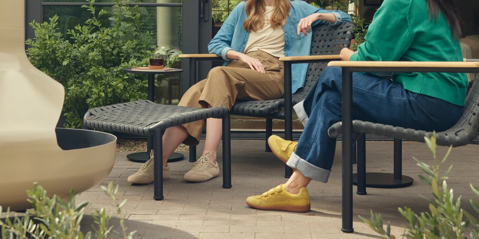 Wide hero banner of two friends in conversation seated on Neighbor Up-Down Chairs with charcoal woven-strap seats and natural-wood armrests on a brick paver patio, with a Neighbor Up-Down Ottoman between them, a small black side table, and the cream-beige curve of a Neighbor Totem Chiminea in the foreground edge in front of a glass-walled garden building.