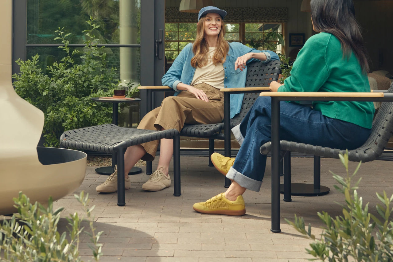 Tablet-crop hero of two friends in conversation seated on Neighbor Up-Down Chairs with charcoal woven-strap seats and natural-wood armrests on a brick paver patio, with an Up-Down Ottoman between them, a small black side table, and the cream-beige curve of a Neighbor Totem Chiminea visible at left in front of a glass-walled garden building.