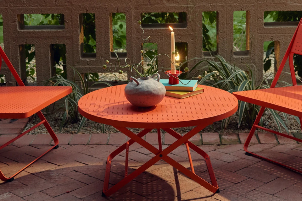 Wide banner of a coral-red Neighbor Take Two Coffee Table on a red-brick paver patio styled with a textured stoneware vase trailing a curling vine, stacked teal and yellow books, and a lit taper candle in a small red holder, with a cinder-block lattice screen wall and tropical greenery in the background.