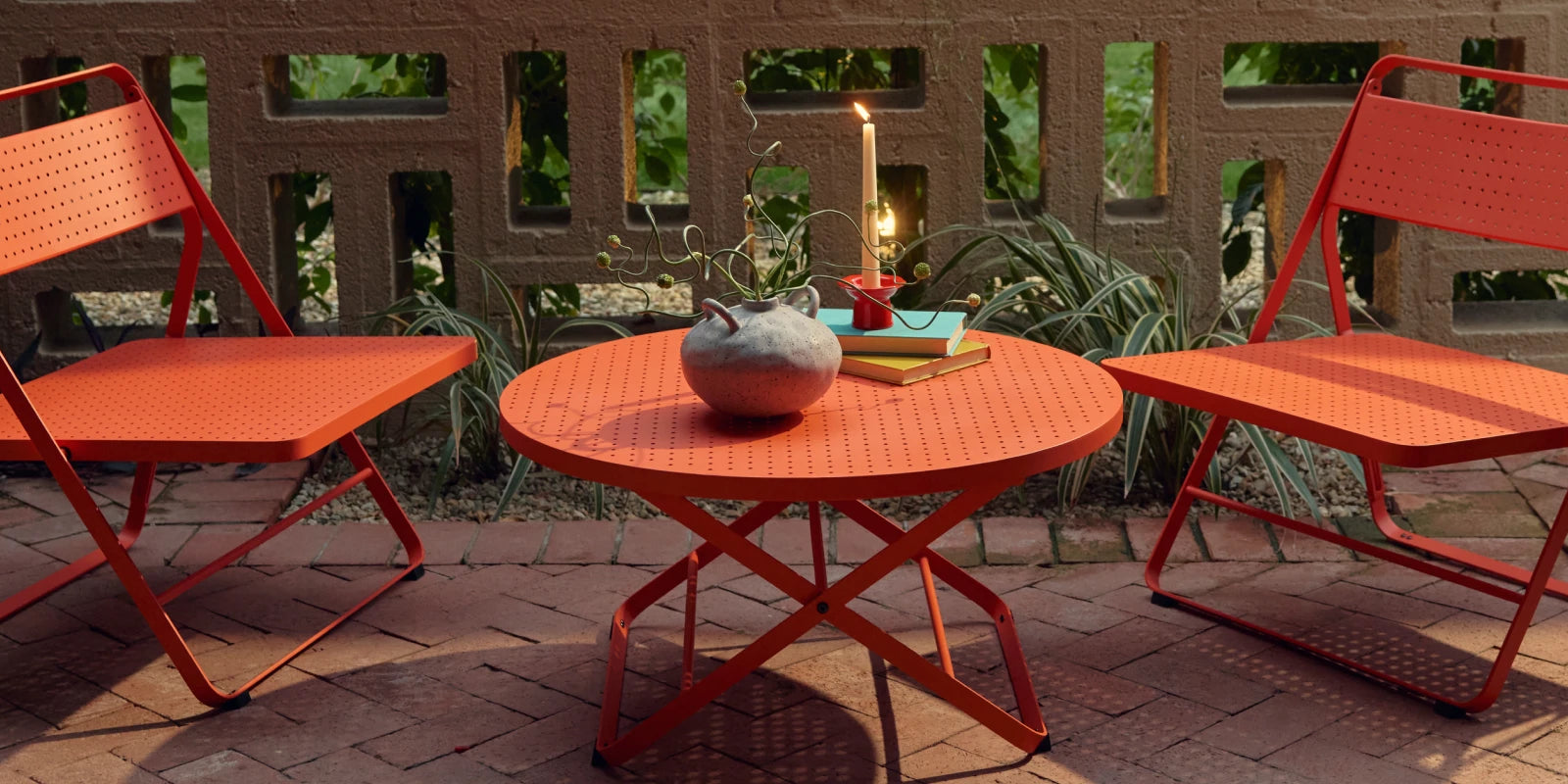 Wide banner of a coral-red Neighbor Take Two Coffee Table on a red-brick paver patio styled with a textured stoneware vase trailing a curling vine, stacked teal and yellow books, and a lit taper candle in a small red holder, with a cinder-block lattice screen wall and tropical greenery in the background.