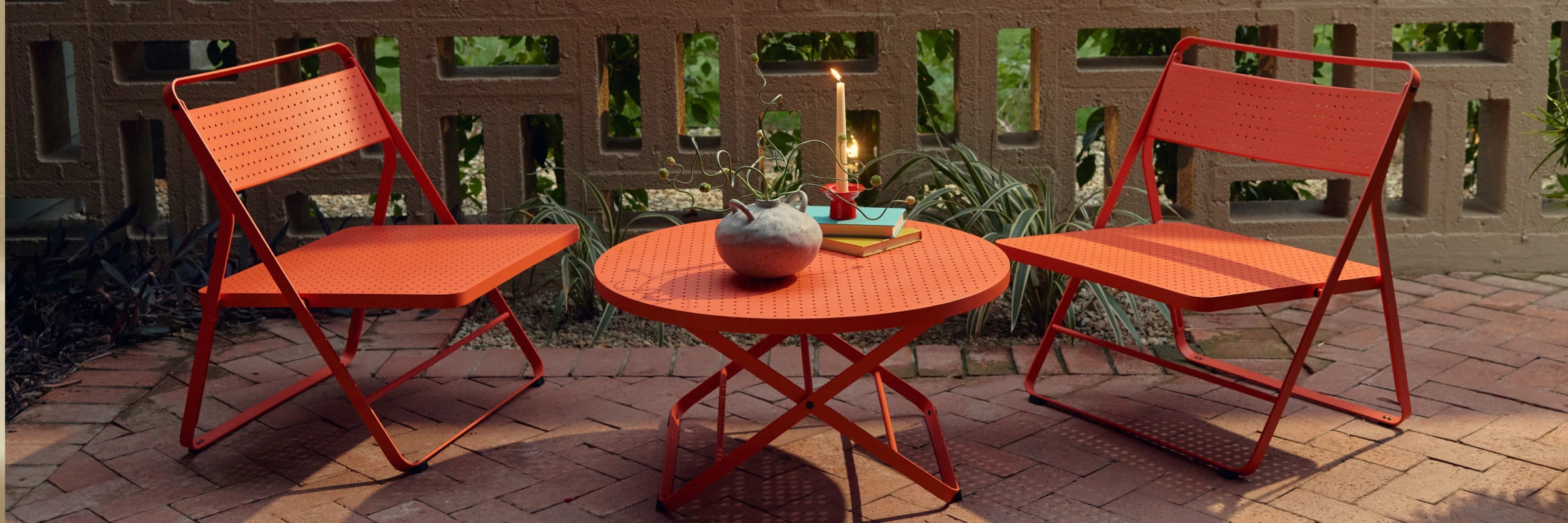 Wide banner of a coral-red Neighbor Take Two Coffee Table on a red-brick paver patio styled with a textured stoneware vase trailing a curling vine, stacked teal and yellow books, and a lit taper candle in a small red holder, with a cinder-block lattice screen wall and tropical greenery in the background.