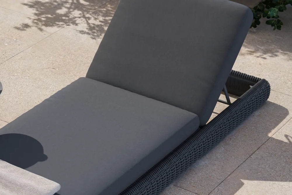 Three-quarter view of a Neighbor Roundabout Sun Lounger in dark charcoal resin wicker with thick charcoal-gray all-weather cushion on a sunlit stone-tile patio, backrest tilted to a relaxed reading angle, with a small round side table holding a glass of lemon water beside it.