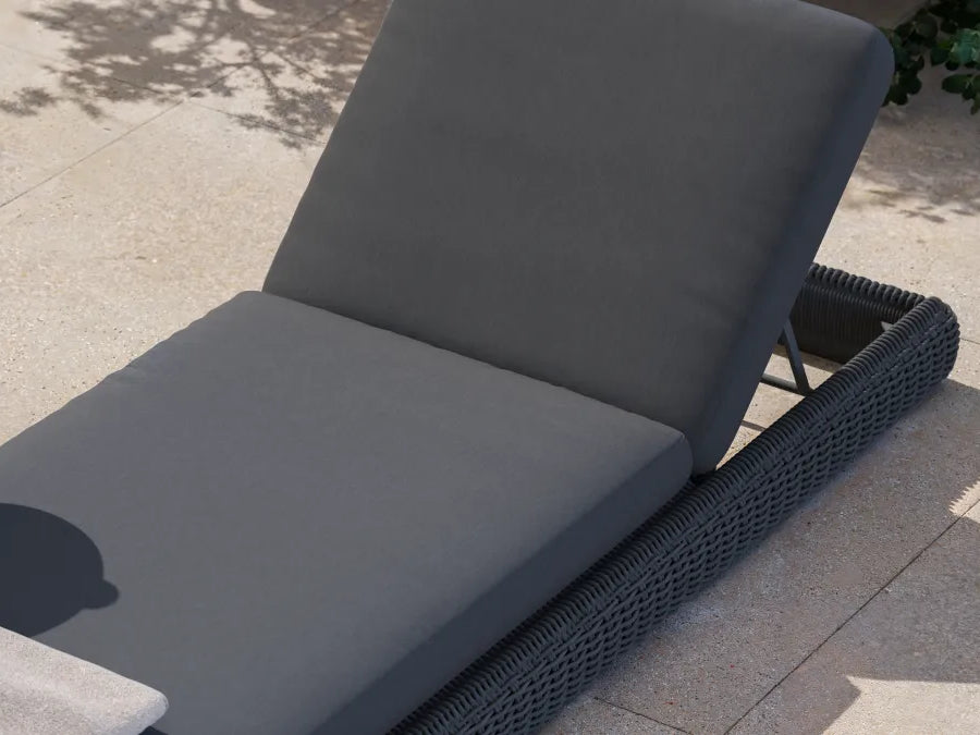 Three-quarter view of a Neighbor Roundabout Sun Lounger in dark charcoal resin wicker with thick charcoal-gray all-weather cushion on a sunlit stone-tile patio, backrest tilted to a relaxed reading angle, with a small round side table holding a glass of lemon water beside it.