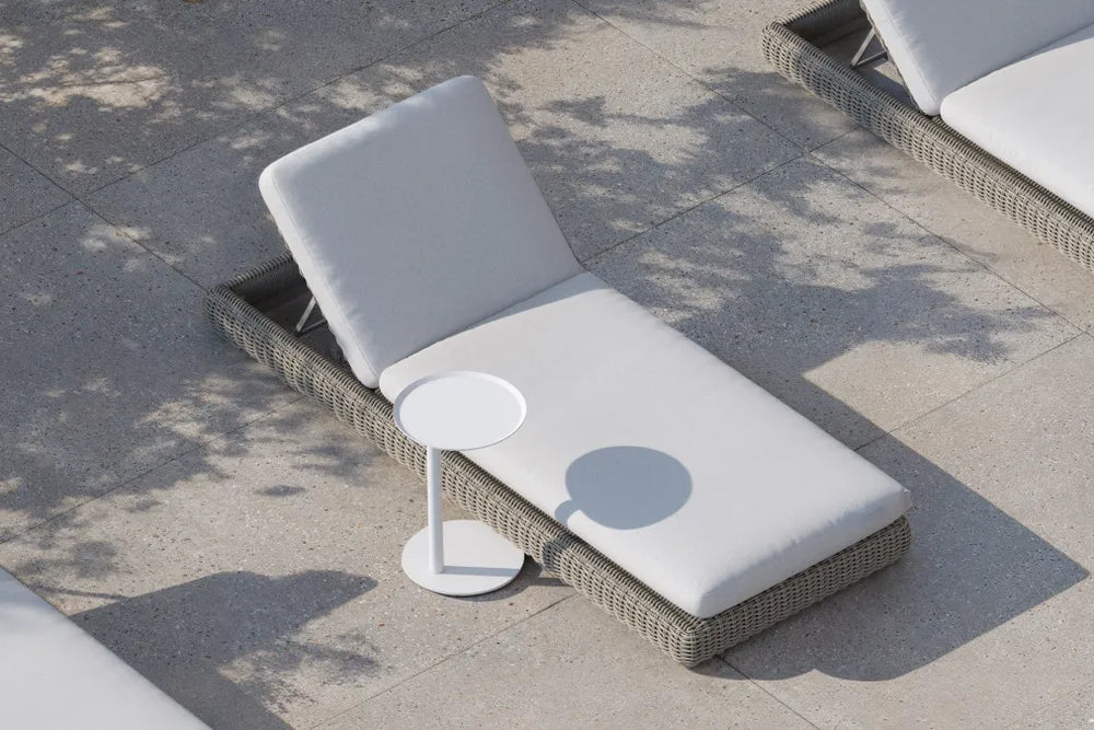 Top-down lifestyle of a Neighbor Roundabout Sun Lounger in taupe-gray resin wicker with cream cushion on a sunlit stone-tile patio, with a small white round side table holding a drink positioned alongside, dappled tree shadows falling across the scene.
