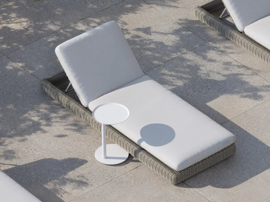 Top-down lifestyle of a Neighbor Roundabout Sun Lounger in taupe-gray resin wicker with cream cushion on a sunlit stone-tile patio, with a small white round side table holding a drink positioned alongside, dappled tree shadows falling across the scene.