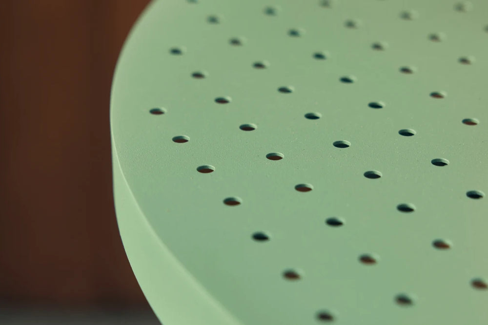Tight macro three-quarter detail of the curved edge of a sage-green Neighbor Take Two Coffee Table top, showing the perforated powder-coated steel and the smooth folded rim that defines the round table, against an out-of-focus stained-wood plank background.
