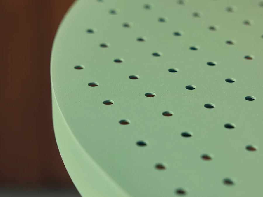 Tight macro three-quarter detail of the curved edge of a sage-green Neighbor Take Two Coffee Table top, showing the perforated powder-coated steel and the smooth folded rim that defines the round table, against an out-of-focus stained-wood plank background.