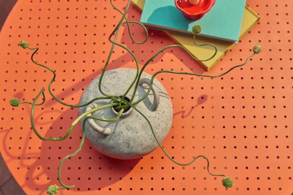 Overhead detail of a coral-orange Neighbor Take Two Coffee Table styled with a textured speckled-stoneware vase trailing a sculptural curling green vine, a stack of teal and yellow hardcover books, and a small red taper candleholder set on top.