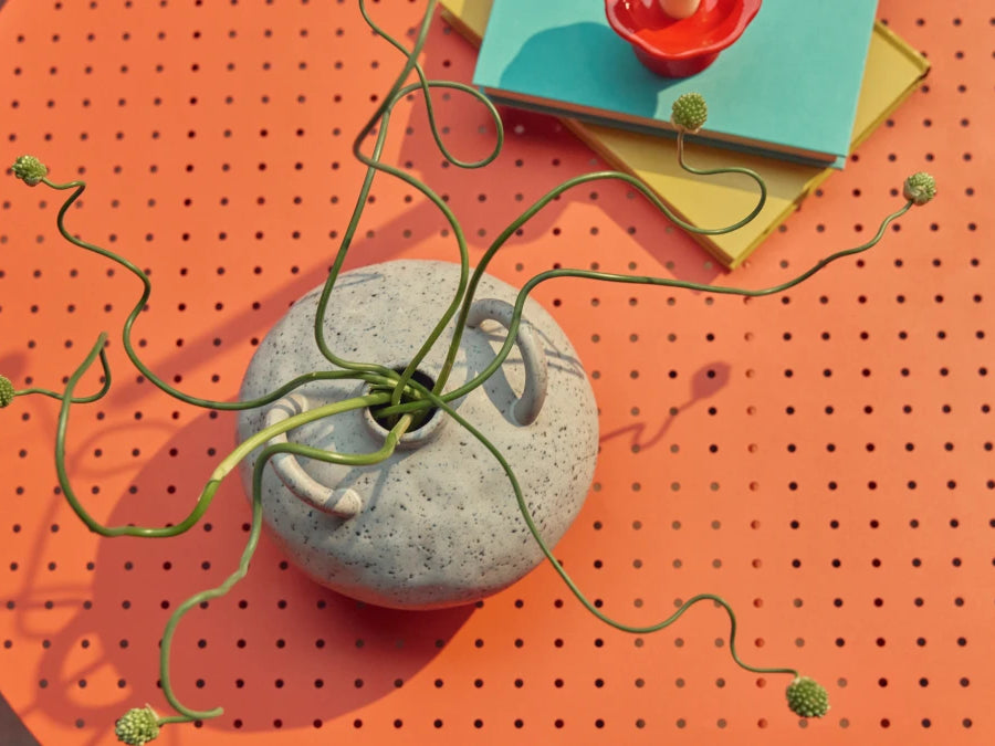 Overhead detail of a coral-orange Neighbor Take Two Coffee Table styled with a textured speckled-stoneware vase trailing a sculptural curling green vine, a stack of teal and yellow hardcover books, and a small red taper candleholder set on top.