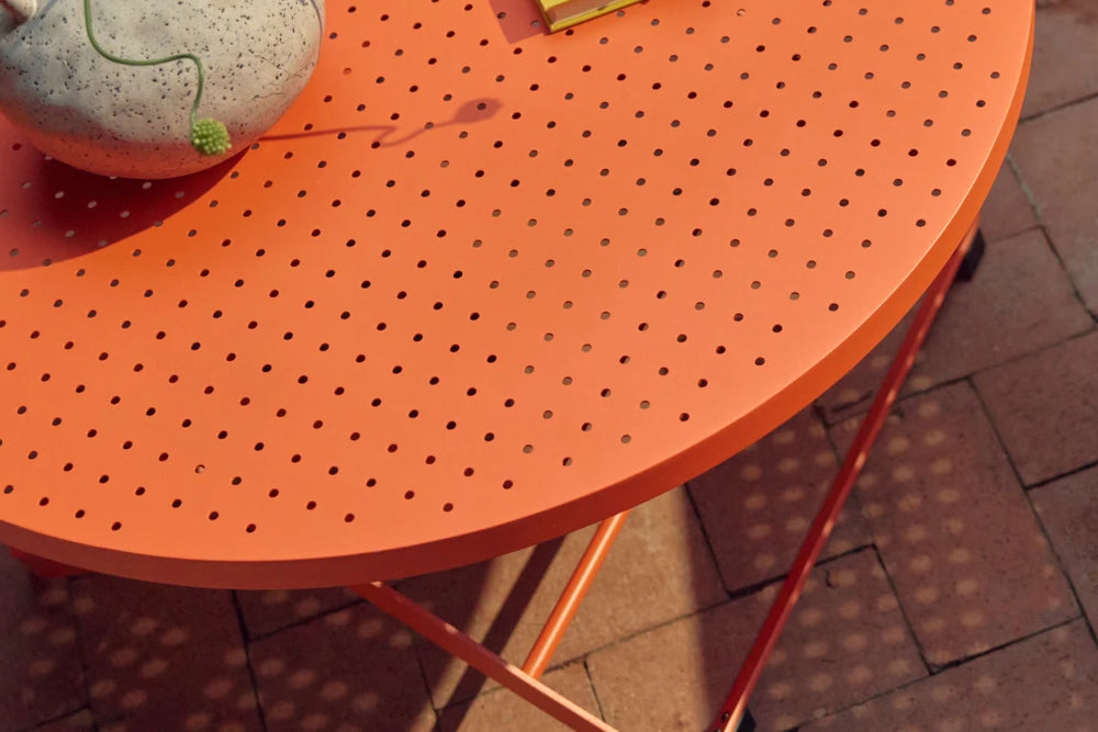 Tight three-quarter detail of the coral-orange Neighbor Take Two Coffee Table top, showing the round perforated steel edge with a speckled stoneware vase on top trailing a curling green vine, casting dappled sunlight through the perforations onto the red-brick paver patio below.