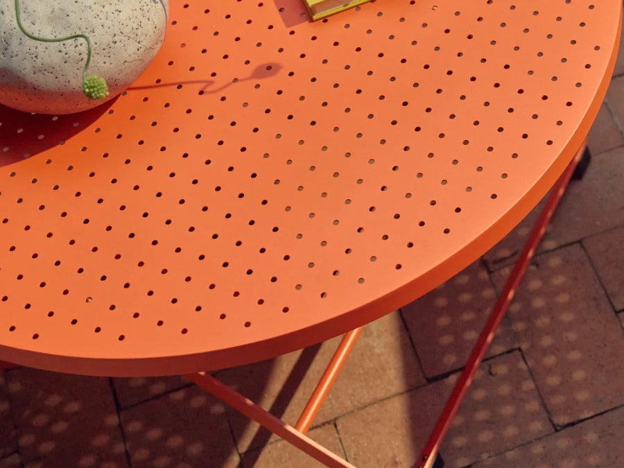 Tight three-quarter detail of the coral-orange Neighbor Take Two Coffee Table top, showing the round perforated steel edge with a speckled stoneware vase on top trailing a curling green vine, casting dappled sunlight through the perforations onto the red-brick paver patio below.