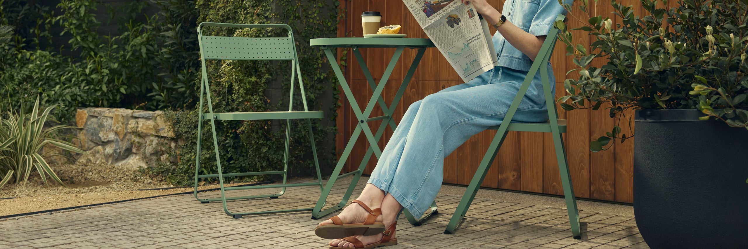 Wide hero crop of a woman in a denim jumpsuit and tan sandals seated reading a newspaper at a sage-green Neighbor Take Two Bistro Set on a cobblestone patio, with a coffee cup and pastry on the round perforated table, an empty matching folding chair across, a stone planter wall, ornamental grasses, and a tall black ceramic planter framing the scene.