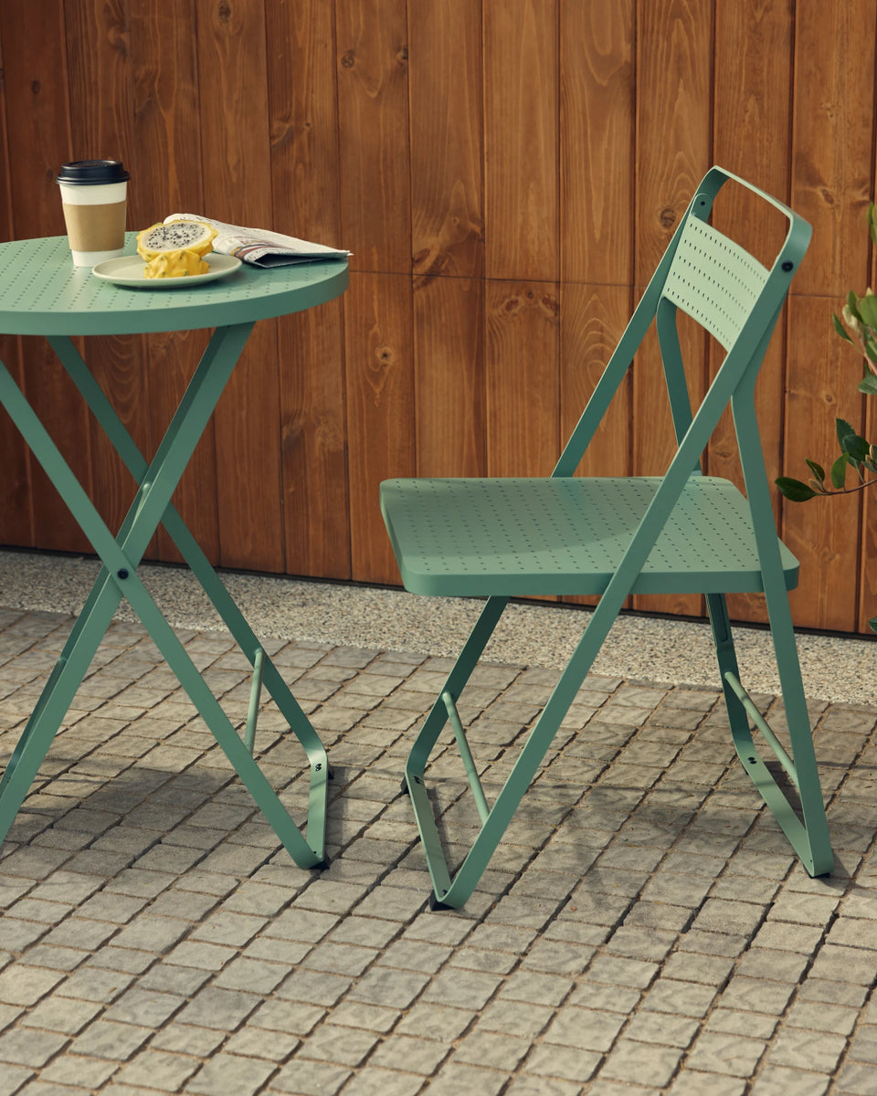 Vertical-crop hero of a sage-green Neighbor Take Two Bistro Table and folding chair on a cobblestone patio against a stained-wood plank wall, with a coffee cup, folded newspaper and small pastry plate on top.