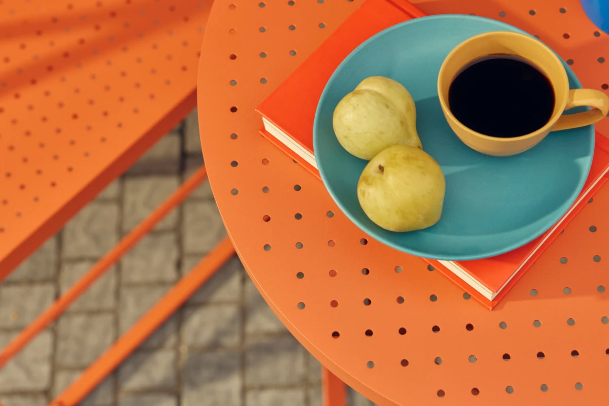 Tight overhead detail of a coral-orange Neighbor Take Two Bistro Table styled with a turquoise plate of yellow pears and a yellow espresso cup of black coffee balanced on a coral-red hardcover book, with a folding chair seat and small blue vase cropped at the edges and a cobblestone patio visible below.