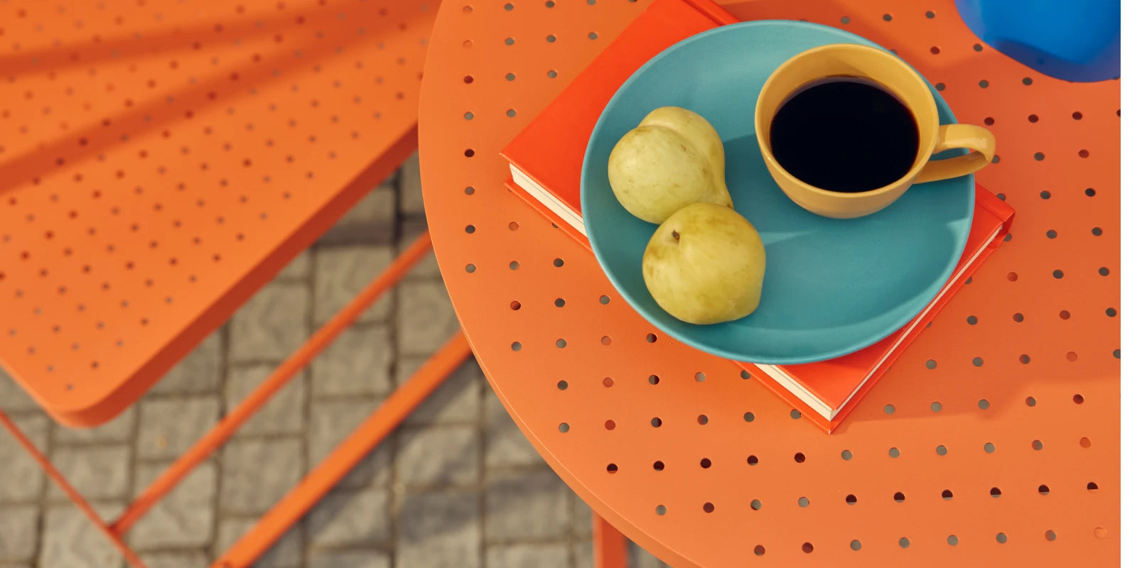Tight overhead detail of a coral-orange Neighbor Take Two Bistro Table styled with a turquoise plate of yellow pears and a yellow espresso cup of black coffee balanced on a coral-red hardcover book, with a folding chair seat and small blue vase cropped at the edges and a cobblestone patio visible below.