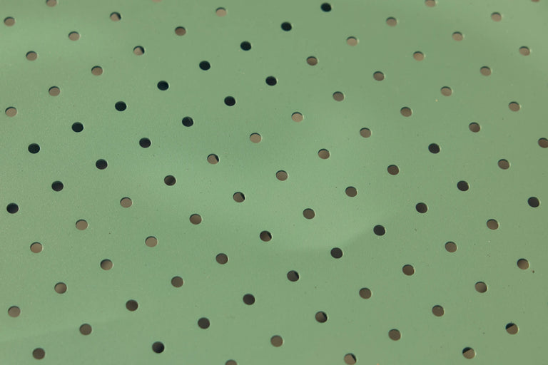 Wide macro of the perforated sage-green powder-coated steel surface of a Neighbor Take Two product, raked sunlight glancing across the round laser-cut holes to highlight the color and texture.