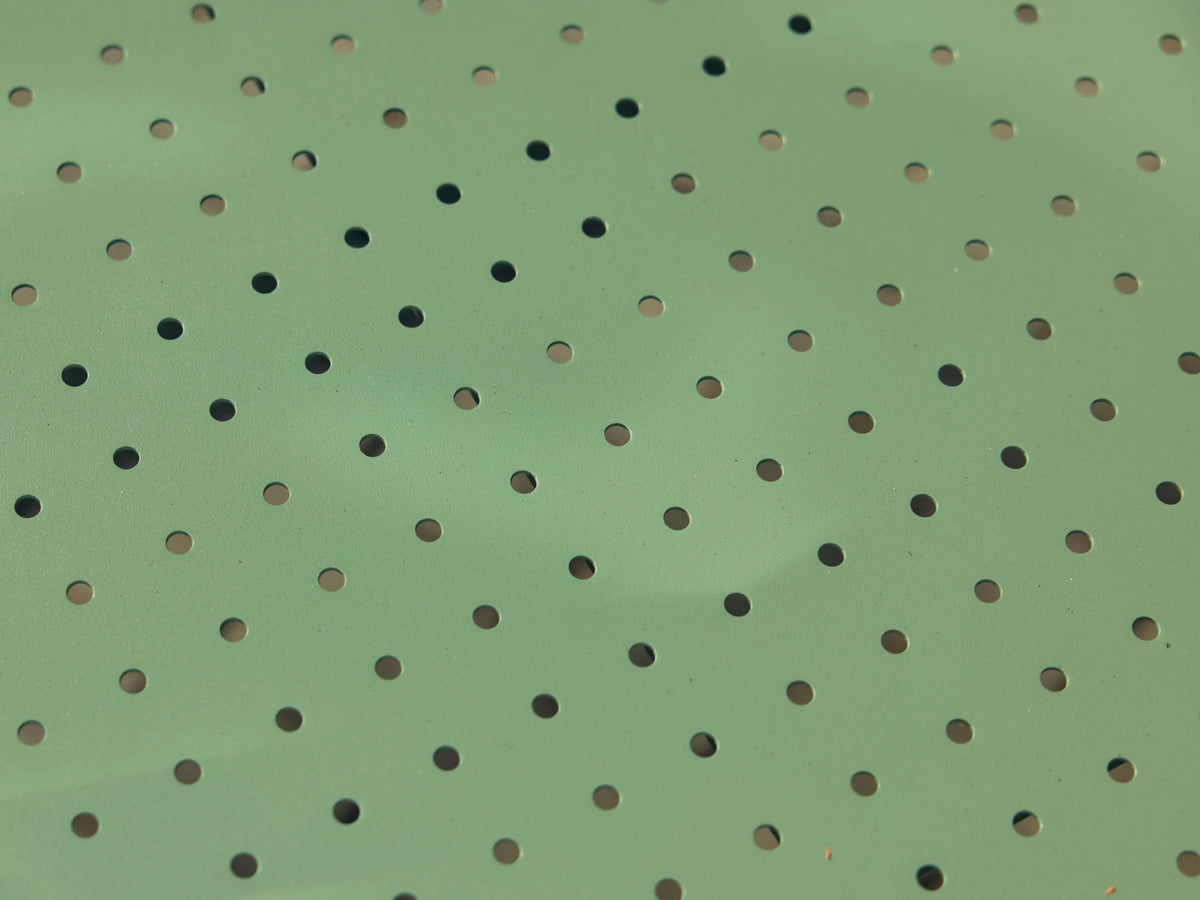 Wide macro of the perforated sage-green powder-coated steel surface of a Neighbor Take Two product, raked sunlight glancing across the round laser-cut holes to highlight the color and texture.