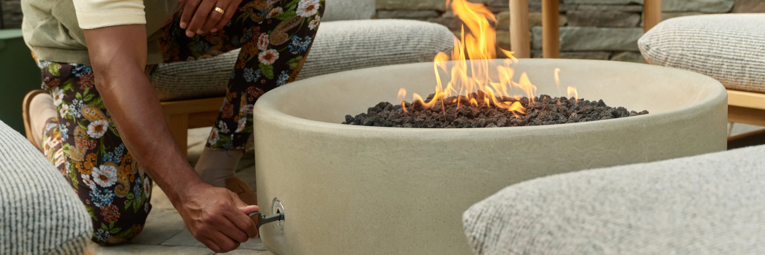 A man in a floral-print outfit crouches beside a cream concrete fire table with a lit flame, with a gray-cushioned teak lounge chair in the foreground on a stone patio