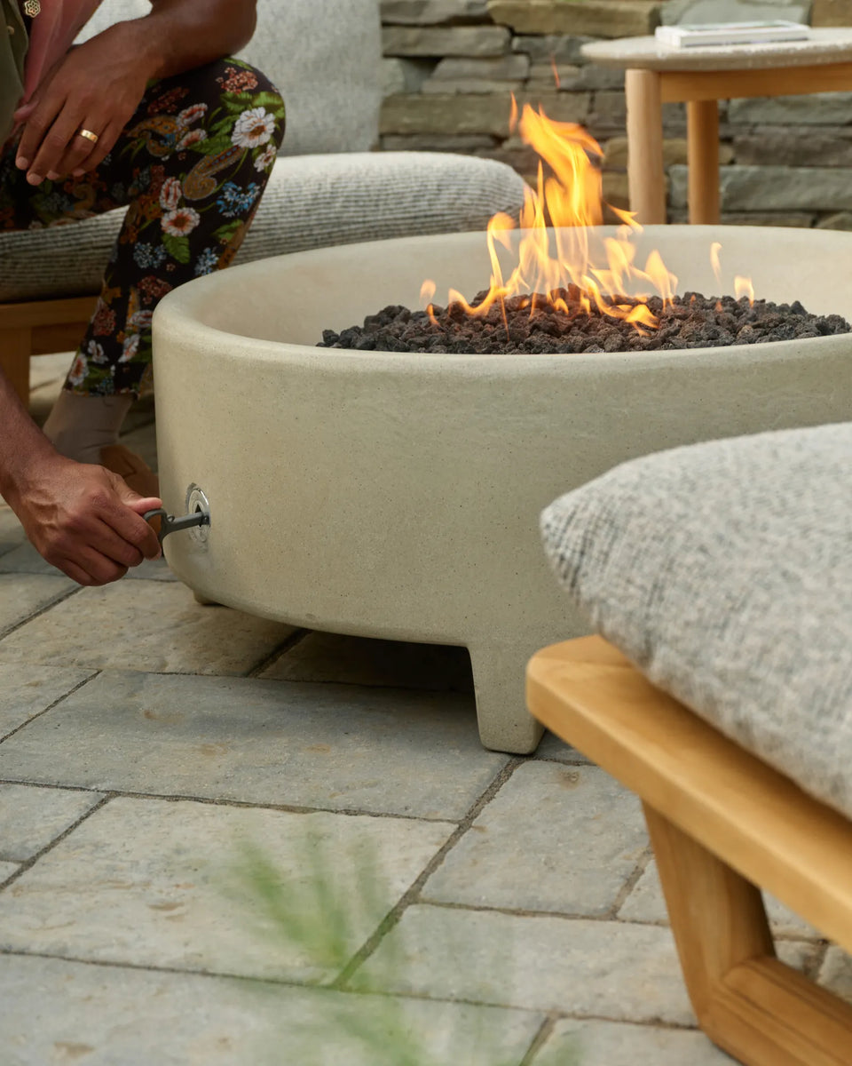 A man in a floral-print outfit crouches beside a cream concrete fire table with a lit flame, with a gray-cushioned teak lounge chair in the foreground on a stone patio