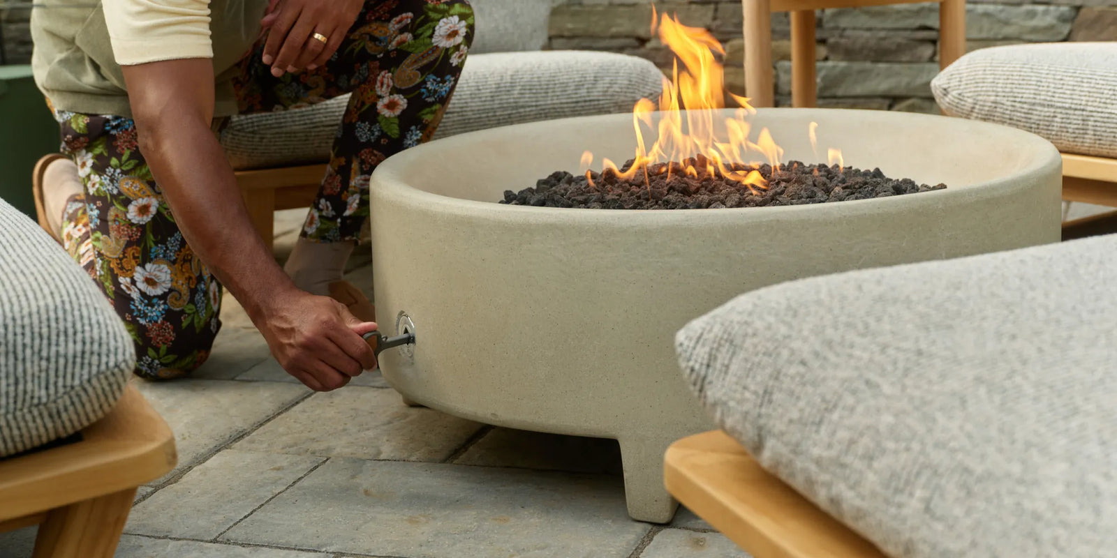 A man in a floral-print outfit crouches beside a cream concrete fire table with a lit flame, with a gray-cushioned teak lounge chair in the foreground on a stone patio