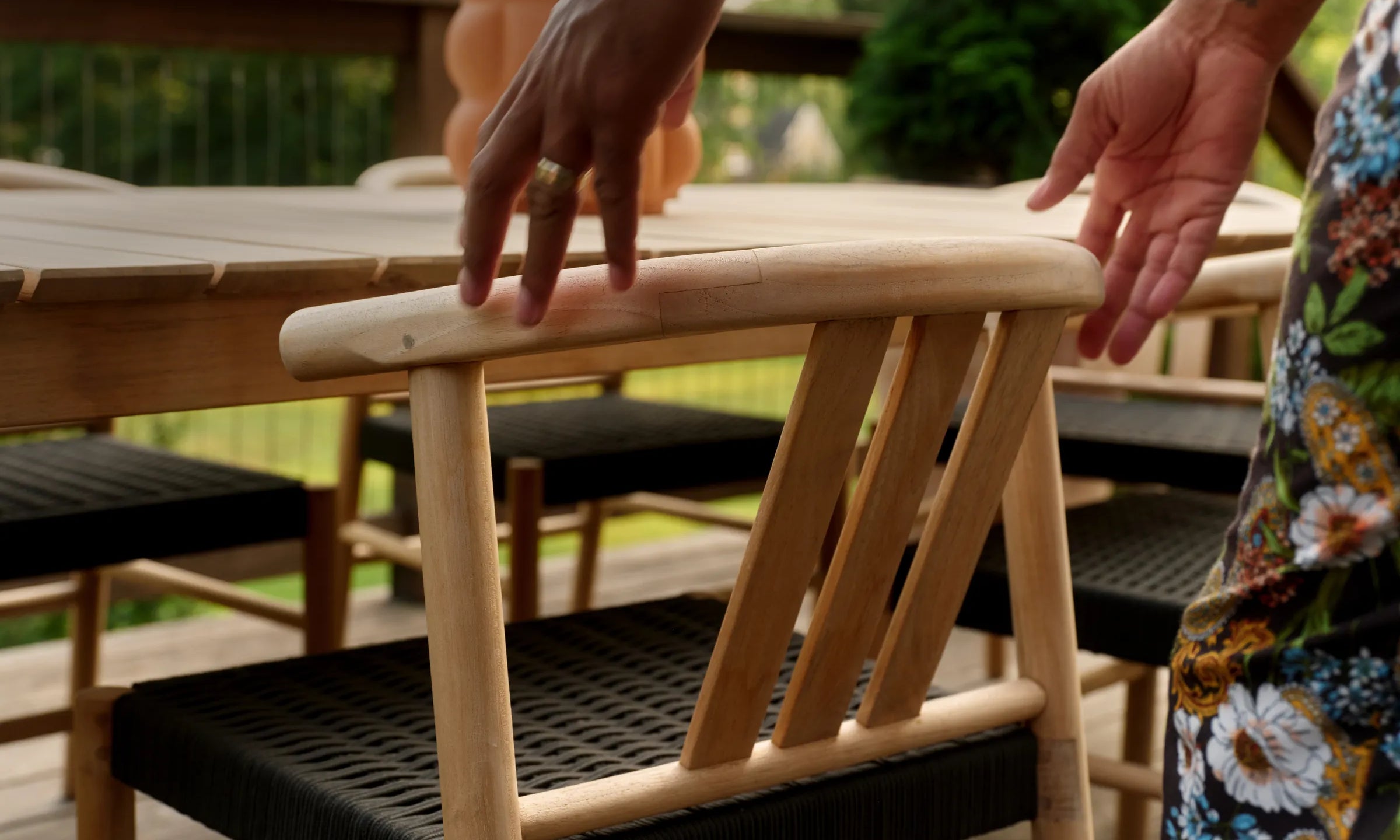A hand rests on the curved top rail of a teak Haven dining chair with a woven black seat, pulled up to a teak dining table on a deck
