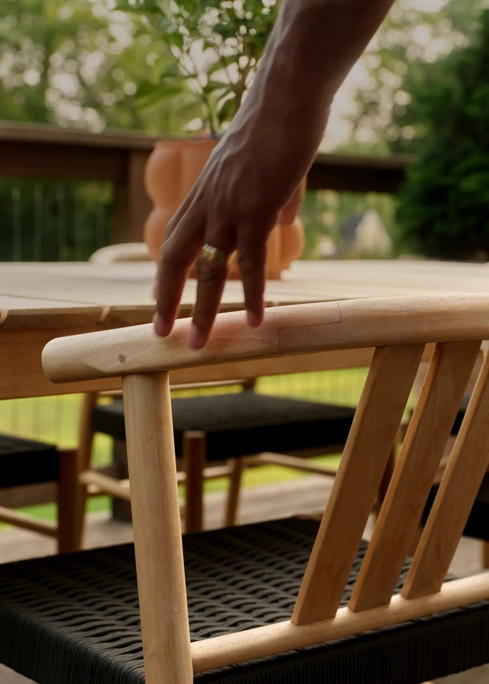 A hand rests on the curved top rail of a teak Haven dining chair with a woven black seat, pulled up to a teak dining table on a deck