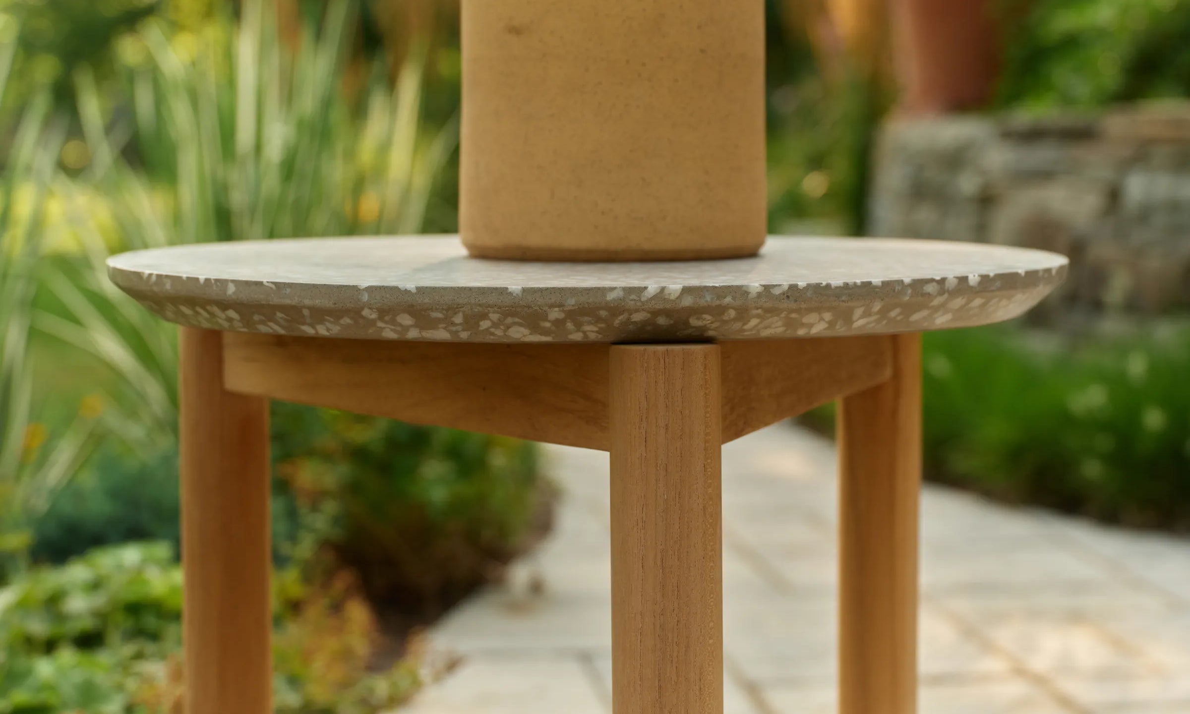 A terracotta planter with a leafy plant sits on a round Haven side table with a stone-look top and teak legs, beside a paved garden path