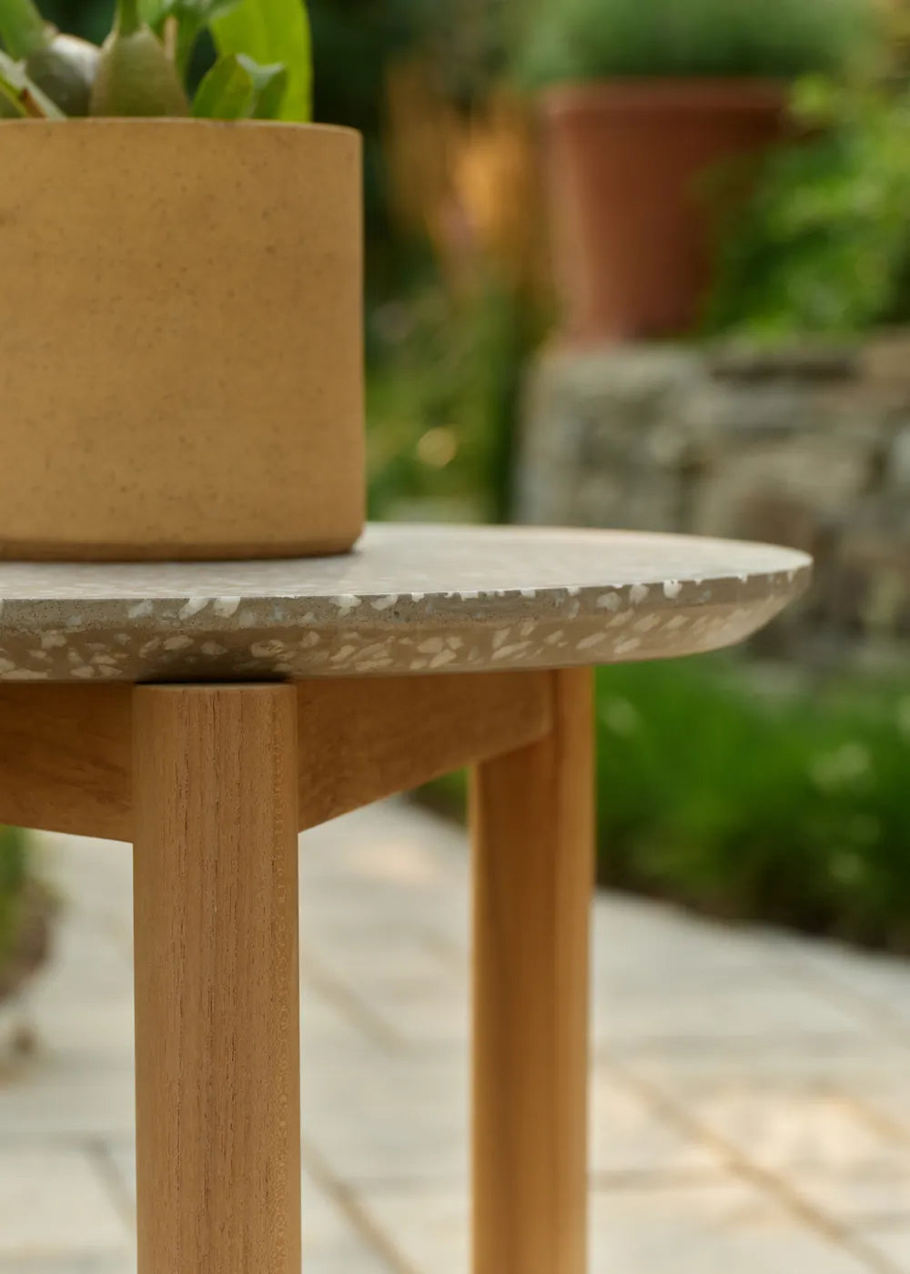 A terracotta planter with a leafy plant sits on a round Haven side table with a stone-look top and teak legs, beside a paved garden path