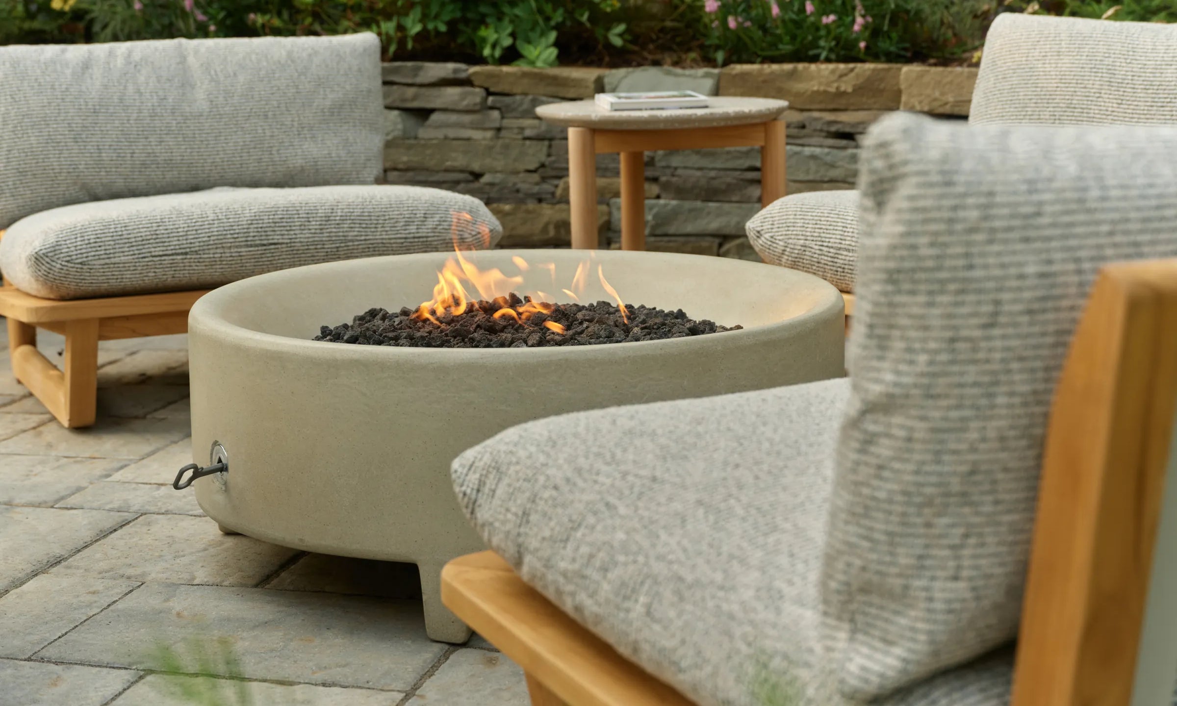 A Rook fire table with a lit flame sits between gray-cushioned teak Dweller lounge chairs on a stone patio, with a small side table in the background