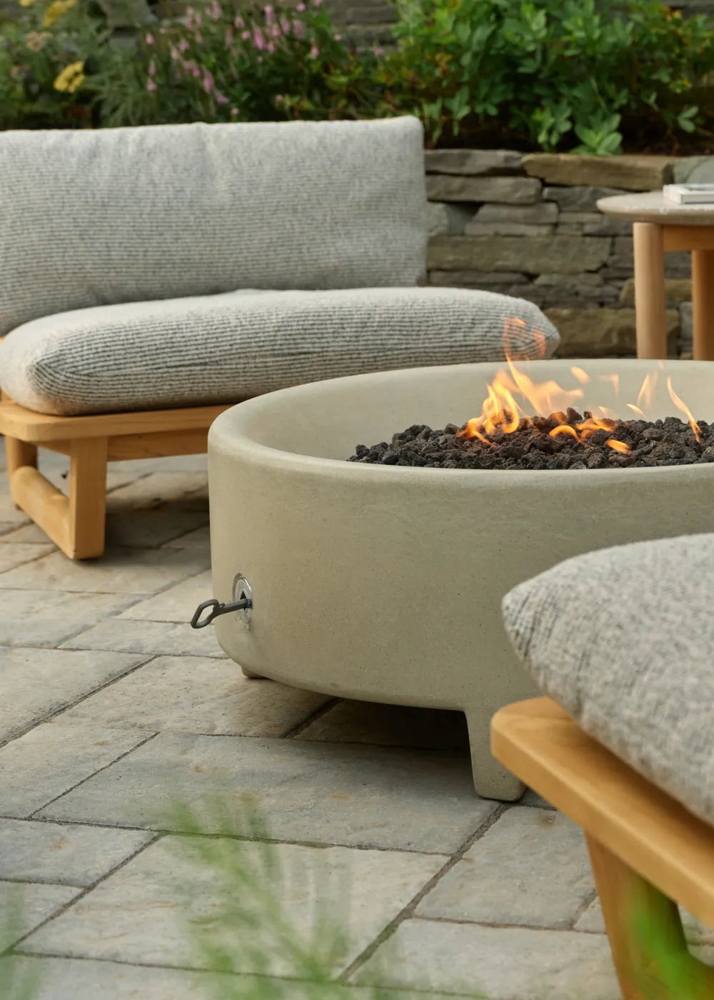 A Rook fire table with a lit flame sits beside a gray-cushioned teak Dweller lounge chair on a stone patio