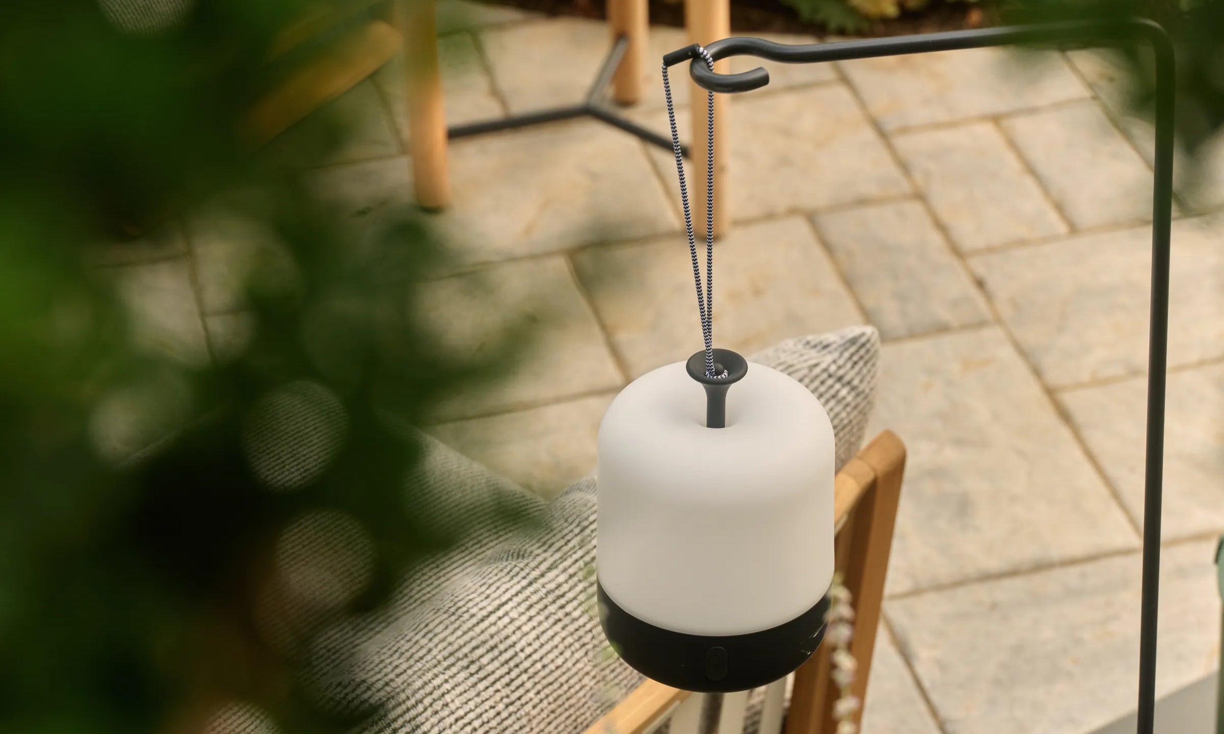 A white Acorn lantern with a black base hangs from a black Acorn lantern stand over a stone patio and a teak chair, framed by blurred foliage