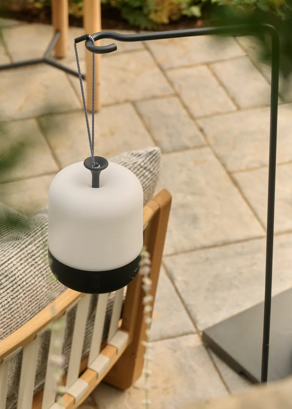 A white Acorn lantern with a black base hangs from a black Acorn lantern stand over a stone patio and a teak chair, framed by blurred foliage