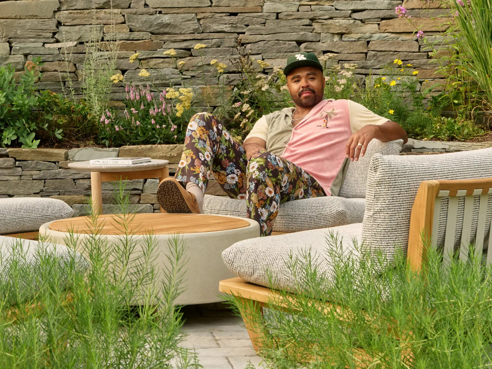 Hilton Carter lounging in a cushioned teak outdoor chair beside a round coffee table in his Baltimore backyard, surrounded by tall grasses, flowering plants, and a stone retaining wall; used as the index banner for the Outside Perspective story Still growing.