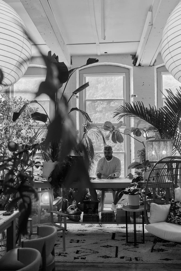 Black-and-white photo of Hilton Carter seated at a table in a bright, plant-filled sunroom, with paper lanterns hanging overhead and tropical plants framing the foreground.