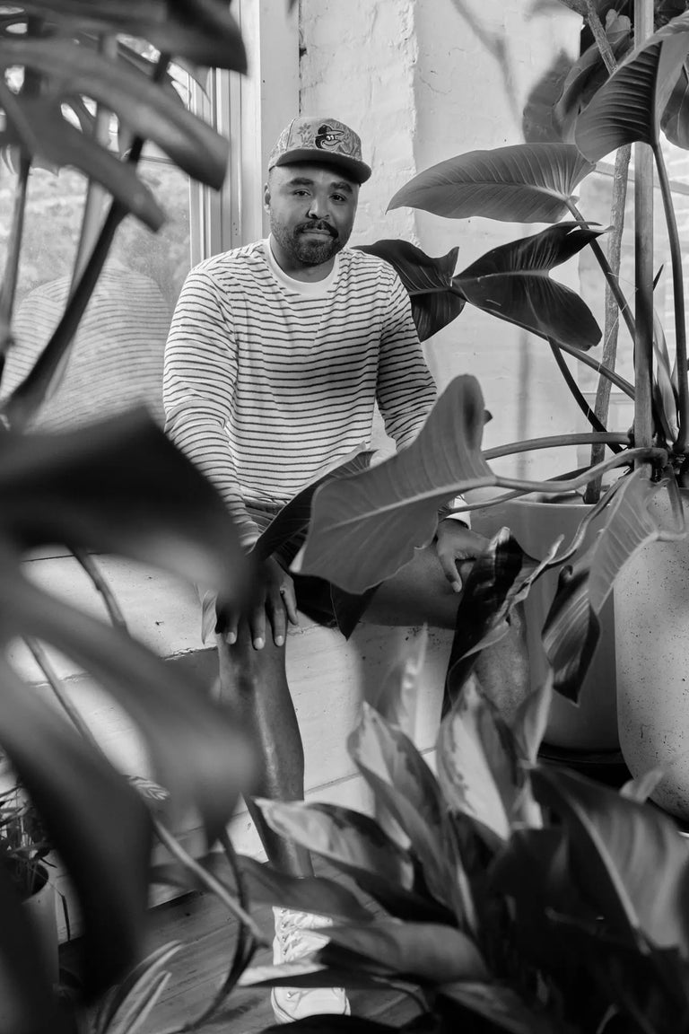 Black-and-white portrait of Hilton Carter in a striped shirt and cap, smiling over the large leaves of a monstera plant.
