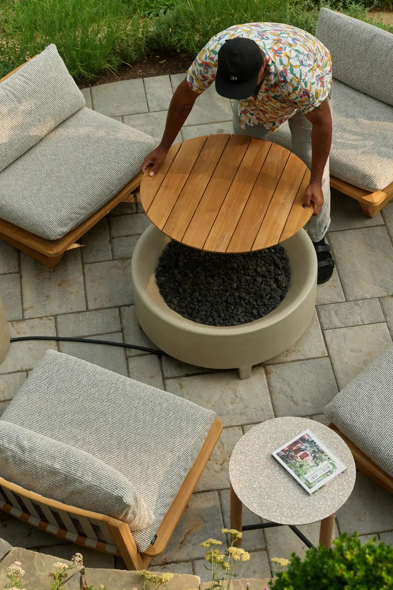 Overhead view of Hilton Carter bending over a round teak coffee table beside a stone fire pit, surrounded by cushioned teak lounge chairs and a small side table on a paved patio.