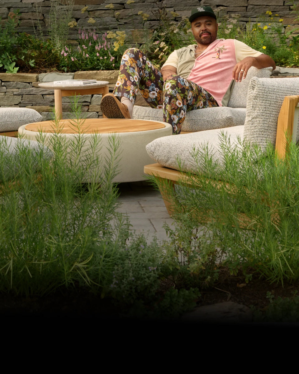 Hilton Carter lounging on a cushioned teak outdoor sectional beside a round coffee table, with tall grasses and a stone retaining wall in his Baltimore backyard.