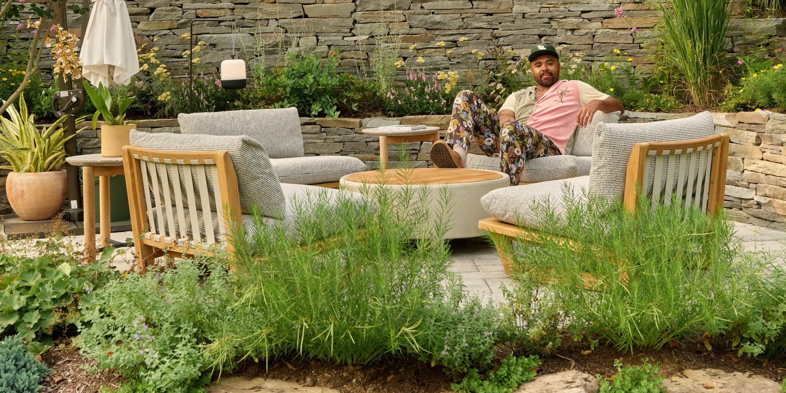 Hilton Carter lounging on a cushioned teak outdoor sectional beside a round coffee table and teak lounge chairs, surrounded by tall grasses, flowering plants, and a stone retaining wall in his Baltimore backyard.