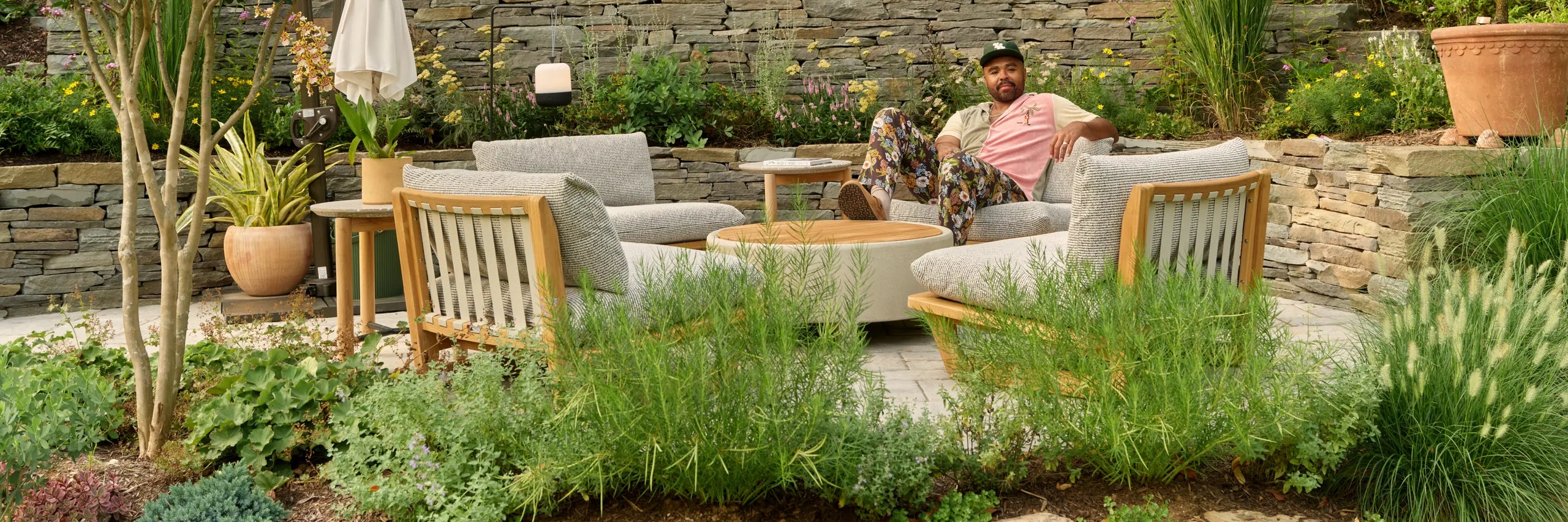 Hilton Carter lounging on a cushioned teak outdoor sectional beside a round coffee table and teak lounge chairs, surrounded by tall grasses, flowering plants, and a stone retaining wall in his Baltimore backyard.