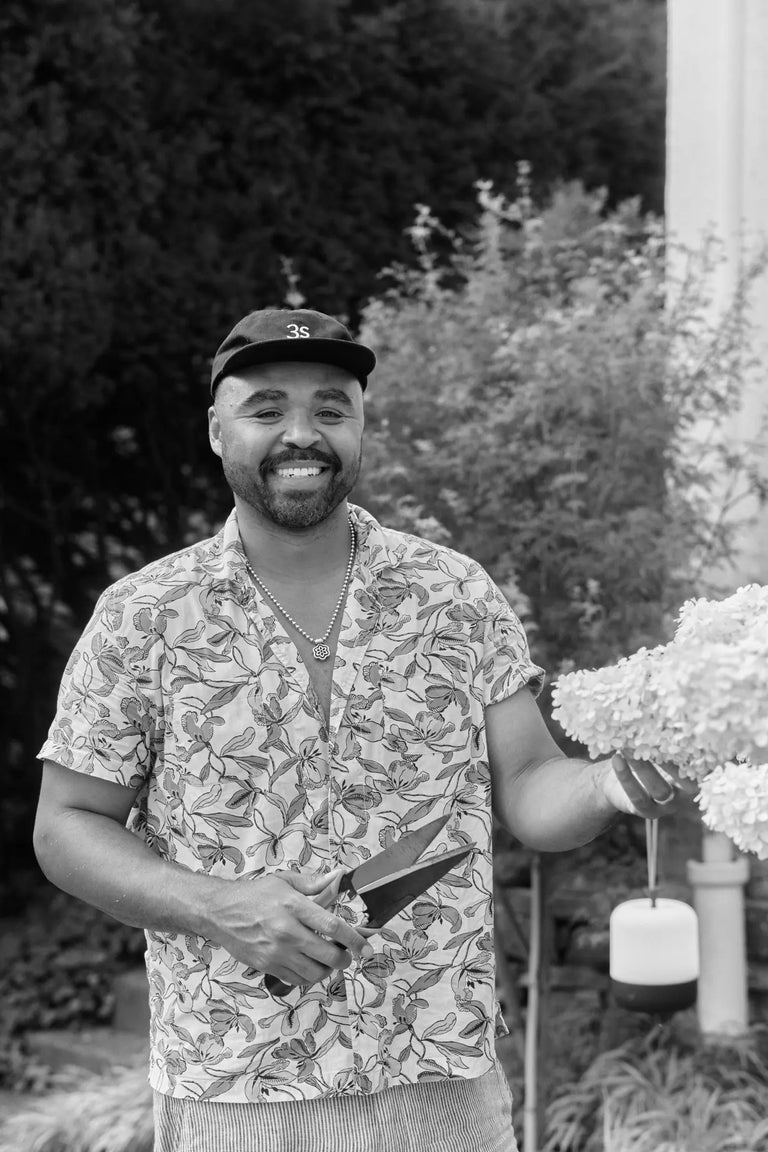 Black-and-white portrait of a smiling Hilton Carter in a cap and patterned short-sleeve shirt, holding pruning shears and a hydrangea bloom in his garden.