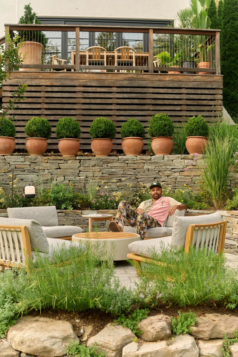 Terraced backyard with a stone retaining wall, teak lounge chairs around a round coffee table, potted boxwoods, and a wooden deck above, with Hilton Carter relaxing in a chair at the right.