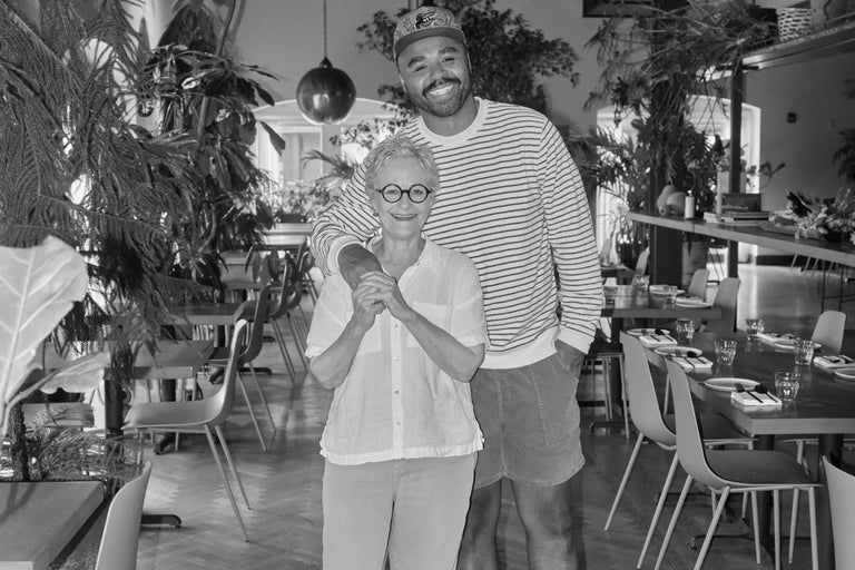 Black-and-white photo of Hilton Carter standing with an older woman in a restaurant dining room with herringbone floors, pendant lights, and chairs.