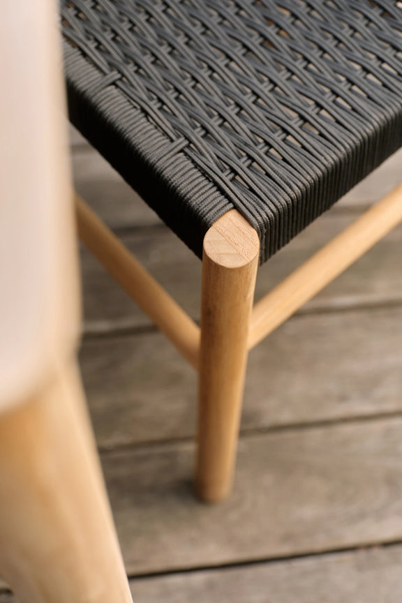 Close-up of a woven black bench seat on a teak frame with a rounded leg, set on a weathered wooden deck.