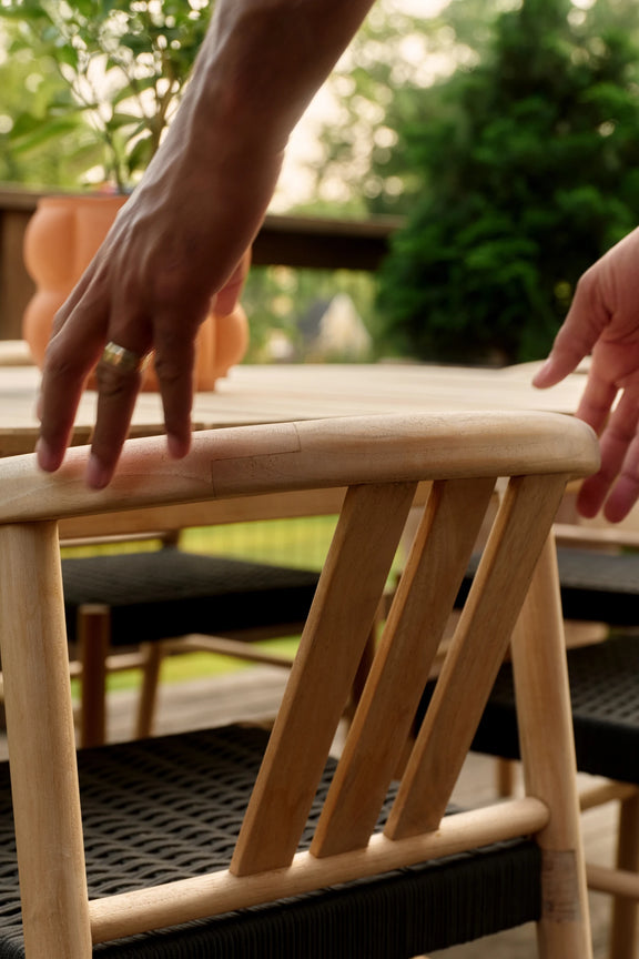 A hand resting on the curved back of a teak dining chair with a woven black seat, with garden greenery blurred behind.